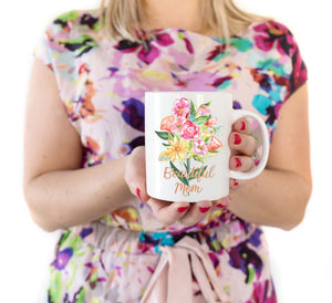 a woman holding a white ceramic mug with a bouquet of hand painted flowers in red, pink, orange, and yellow with green leaves and stems printed on the mug. Below the bouquet the words "Beautiful Mom" in a gold cursive font